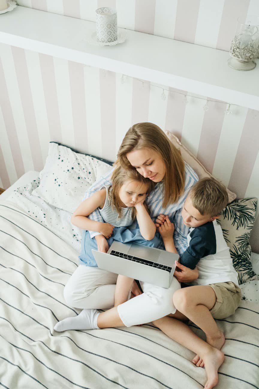 Mother and two children sitting on a bed, looking at a laptop, in a cozy bedroom setting with striped walls and decorative pillows.