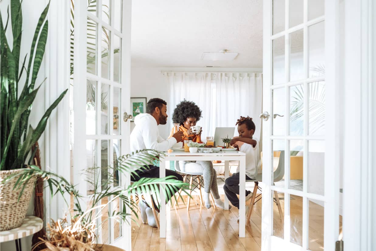 Family enjoying breakfast at a dining table, discussing home buying tips, with a laptop and plants in a bright, airy kitchen setting.