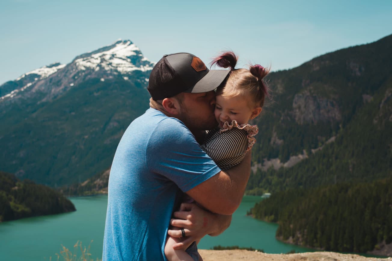 Father holding and kissing daughter against a scenic mountain backdrop with a lake, emphasizing family connection and outdoor adventure.
