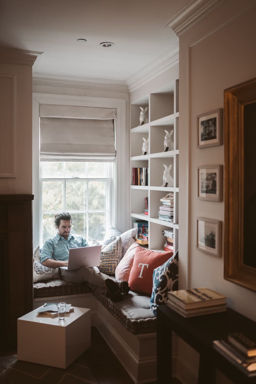 Man sitting in a cozy window nook using a laptop, surrounded by bookshelves and decorative pillows, illustrating a comfortable home environment relevant to first-time home buyers and property valuation discussions.